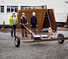 Tre personer og en hund står ved en tilhenger på grus foran en bygning.